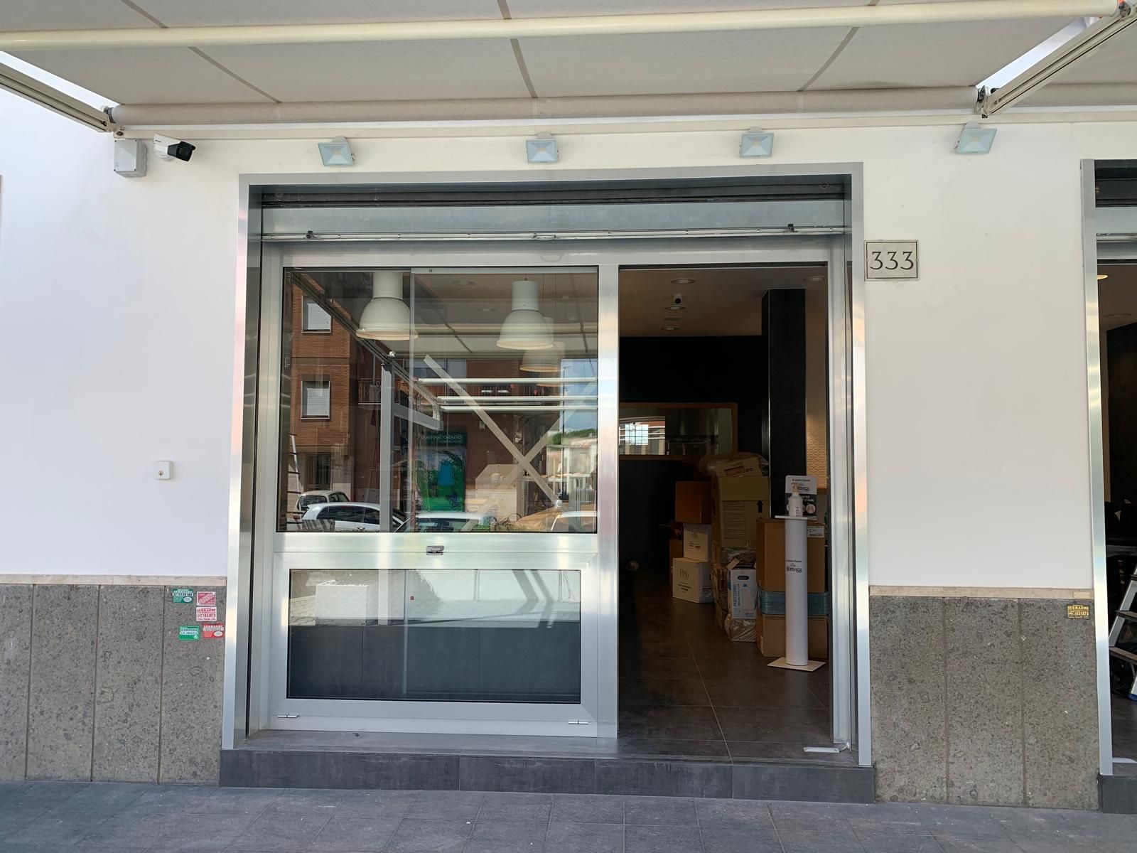 Ingresso del negozio con porta scorrevole in vetro, porta aperta e facciata esterna bianca sotto una tenda da sole