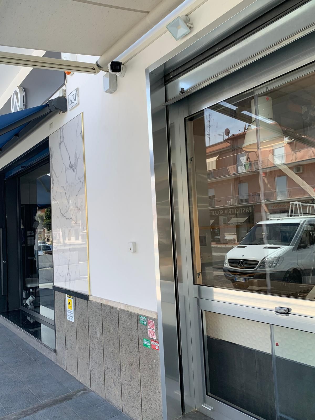 Vetrina con finestra in vetro e parete bianca affacciata sul marciapiede, che riflette gli edifici circostanti e un'auto parcheggiata.