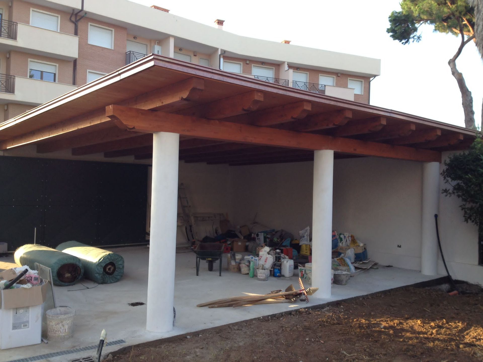 Posto auto coperto esterno con tetto in legno, colonne bianche e vani portaoggetti sottostanti, adiacente a un'abitazione.