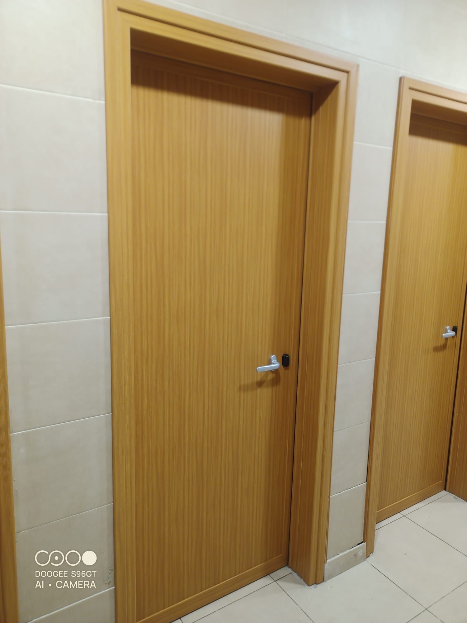 Due porte di legno che conducono a un bagno pubblico, in un corridoio piastrellato di beige, una delle quali parzialmente visibile sulla destra.