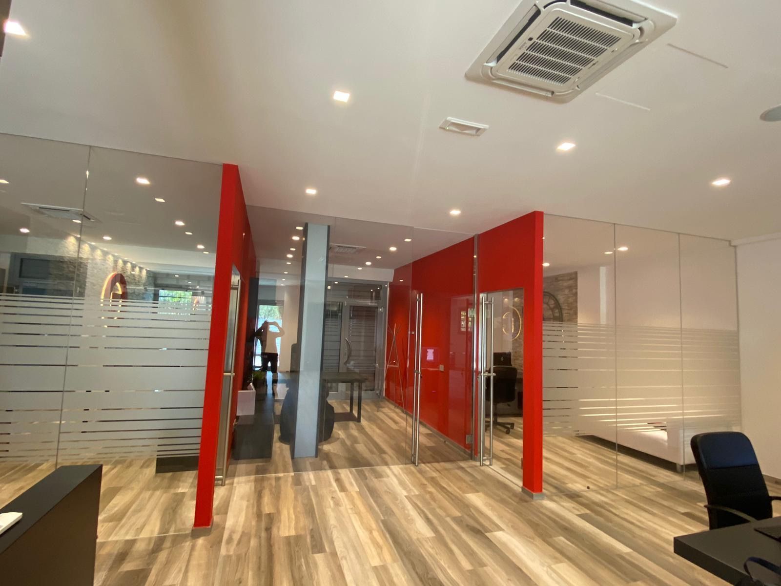Corridoio di un ufficio moderno con pareti divisorie in vetro rosso, porte satinate, pavimento in legno e faretti a incasso a soffitto.
