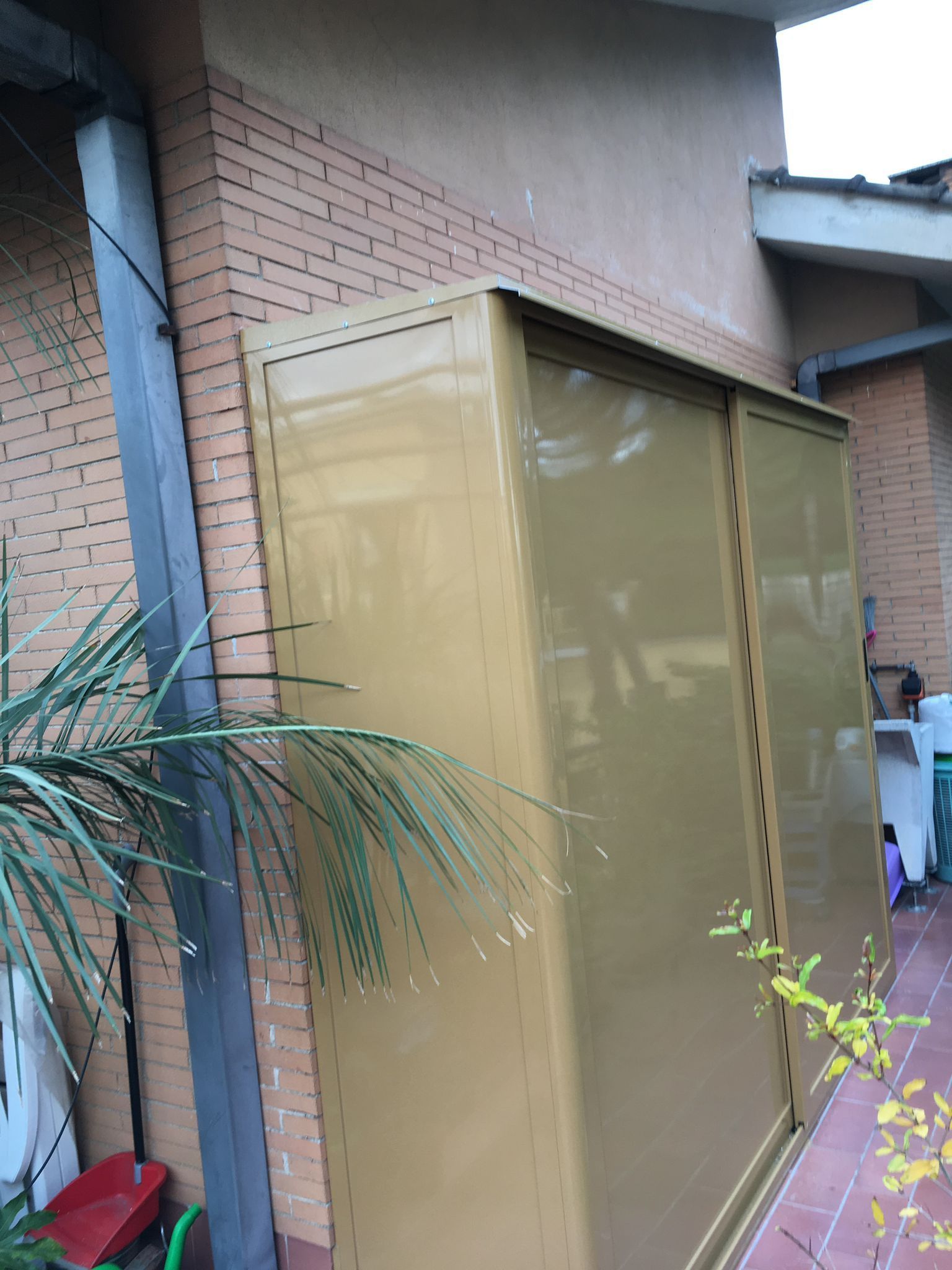 Capanno in metallo color beige accanto a una casa in mattoni, con piante in primo piano.