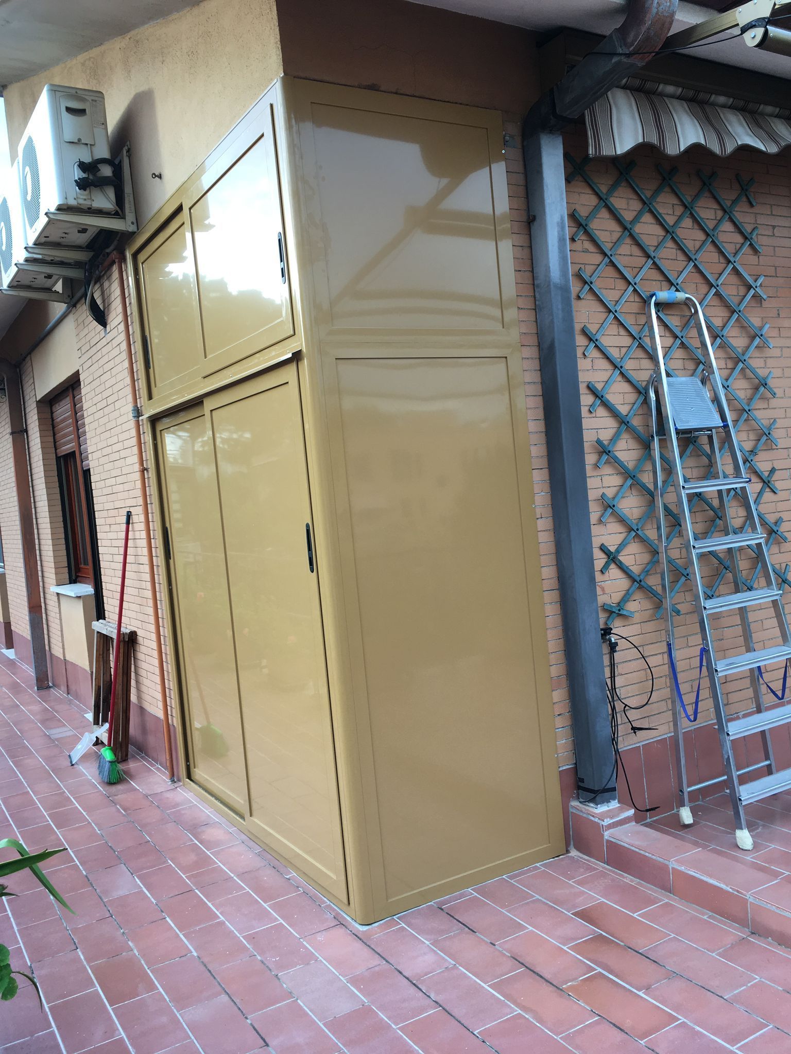 Armadio contenitore in metallo beige accanto a un vialetto in mattoni e a un cancello a rete metallica sulla facciata di un edificio.
