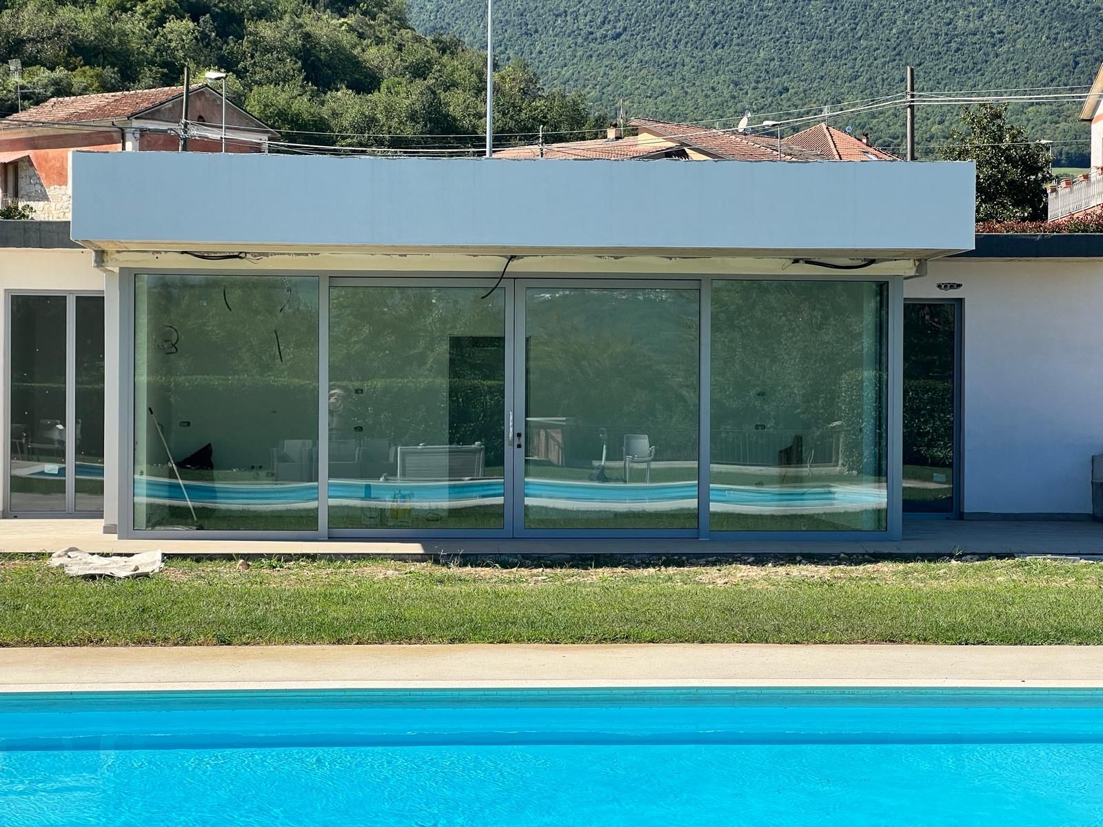 Casa moderna con facciata in vetro, situata accanto a una piscina turchese, con le montagne sullo sfondo.