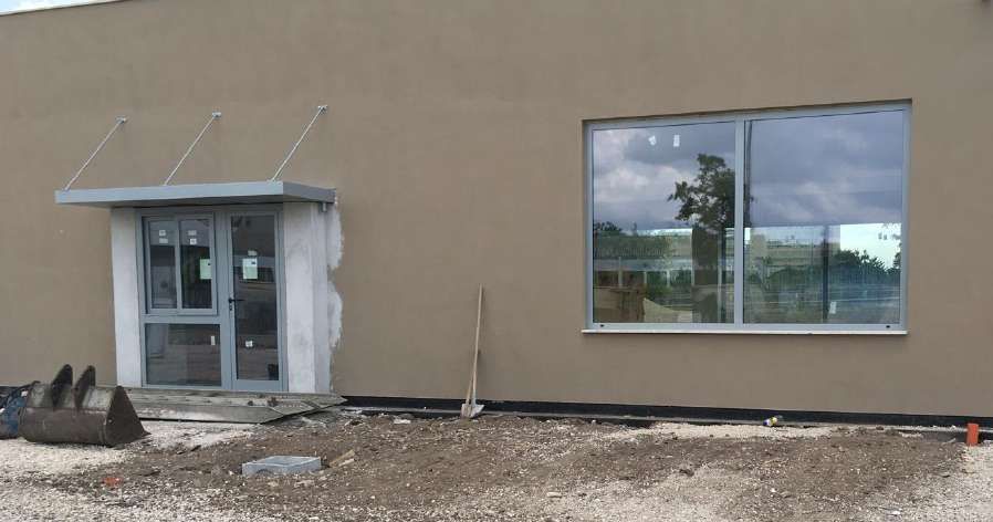 Esterno dell'edificio con porta grigia, ampia finestra e area di cantiere in ghiaia antistante.