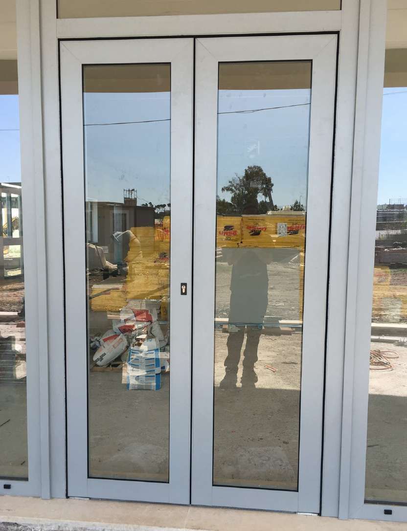 Doppie porte a vetri bianche all'ingresso di un edificio, che riflettono un cantiere edile all'esterno.