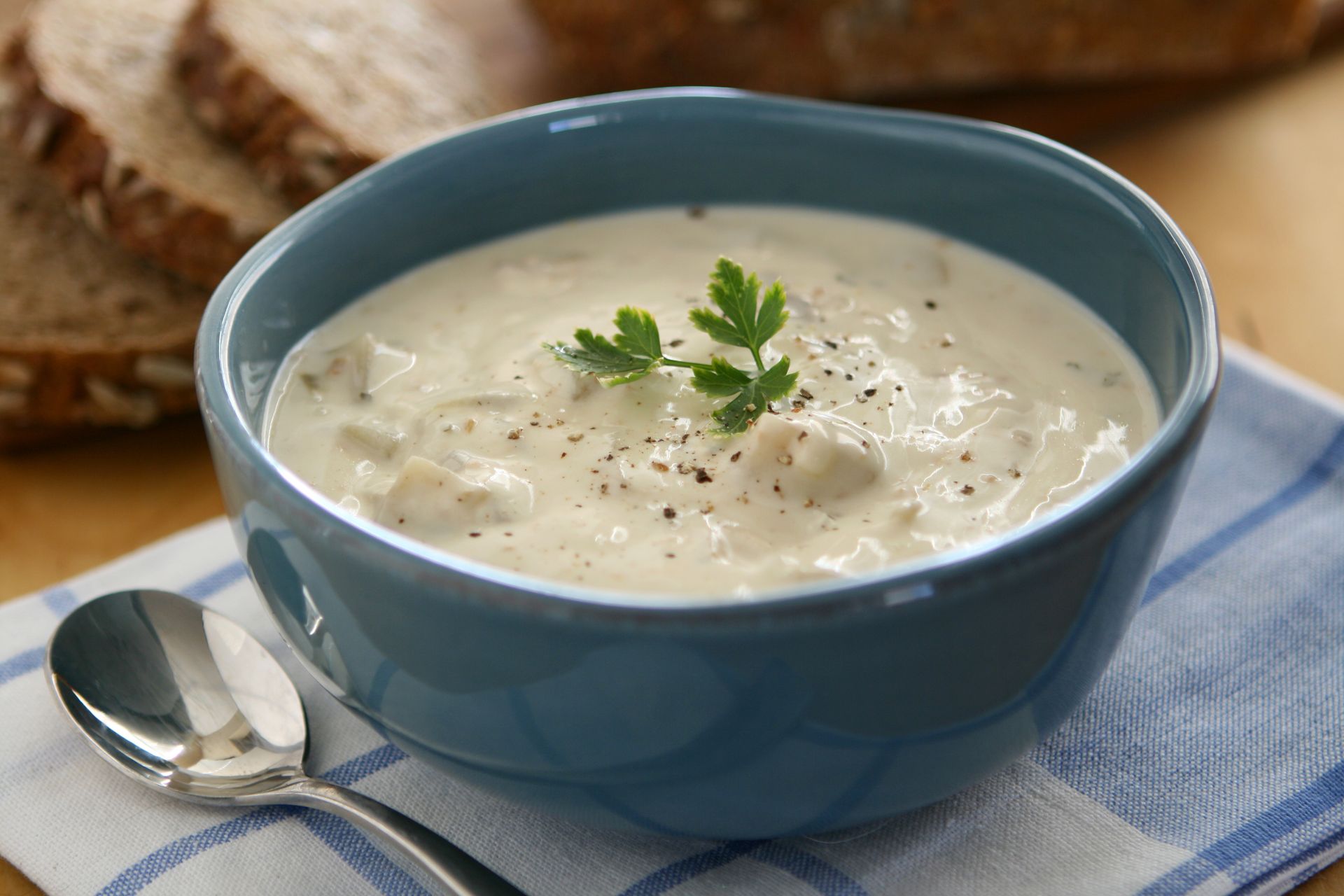 Creamy white soup in a blue bowl with parsley garnish, spoon, and bread slices on a striped cloth.
