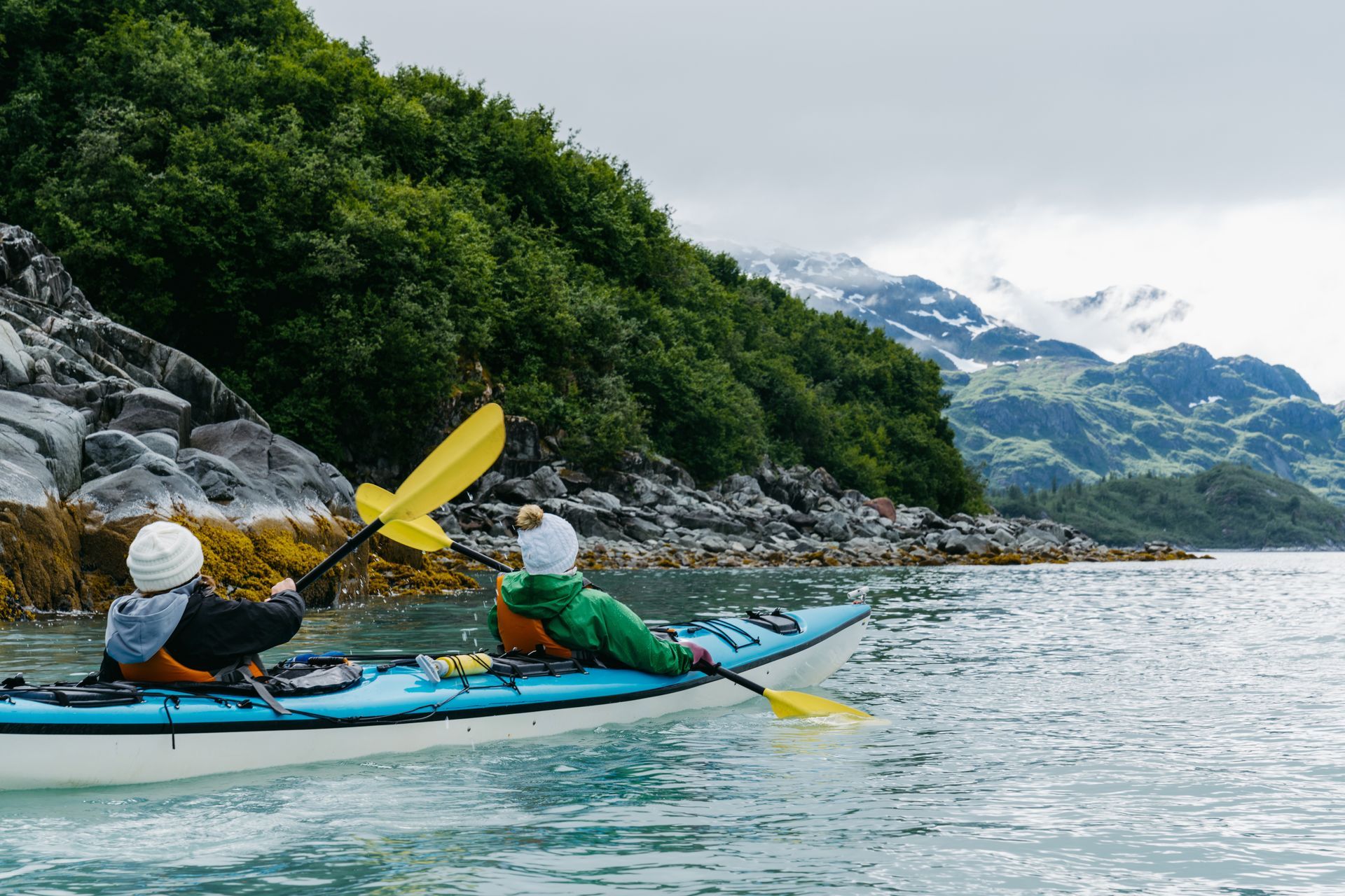 Two people kayaking on a blue kayak, near a rocky shoreline and mountains.
