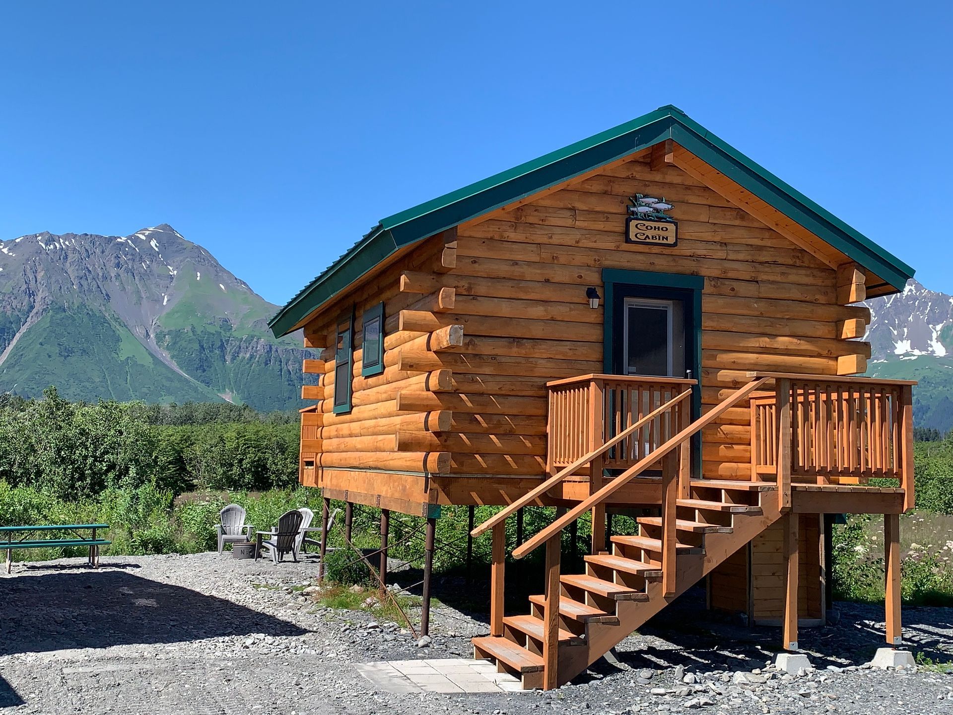 Wooden cabin with stairs, door, and green roof, mountains and blue sky in the background.