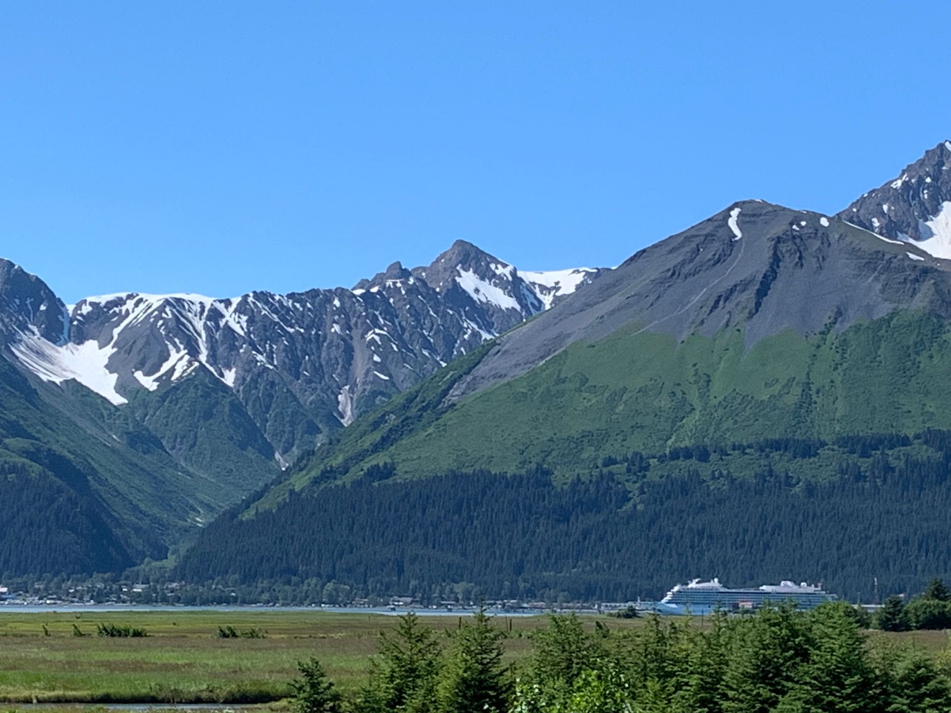 Mountains with snowy peaks overlooking a harbor, with a cruise ship and green fields. Blue sky.