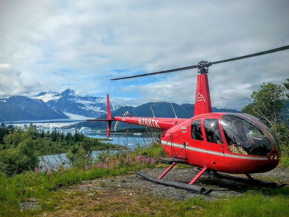 Two red and white helicopters flying near a mountainous, tree-covered cliff under a cloudy sky.