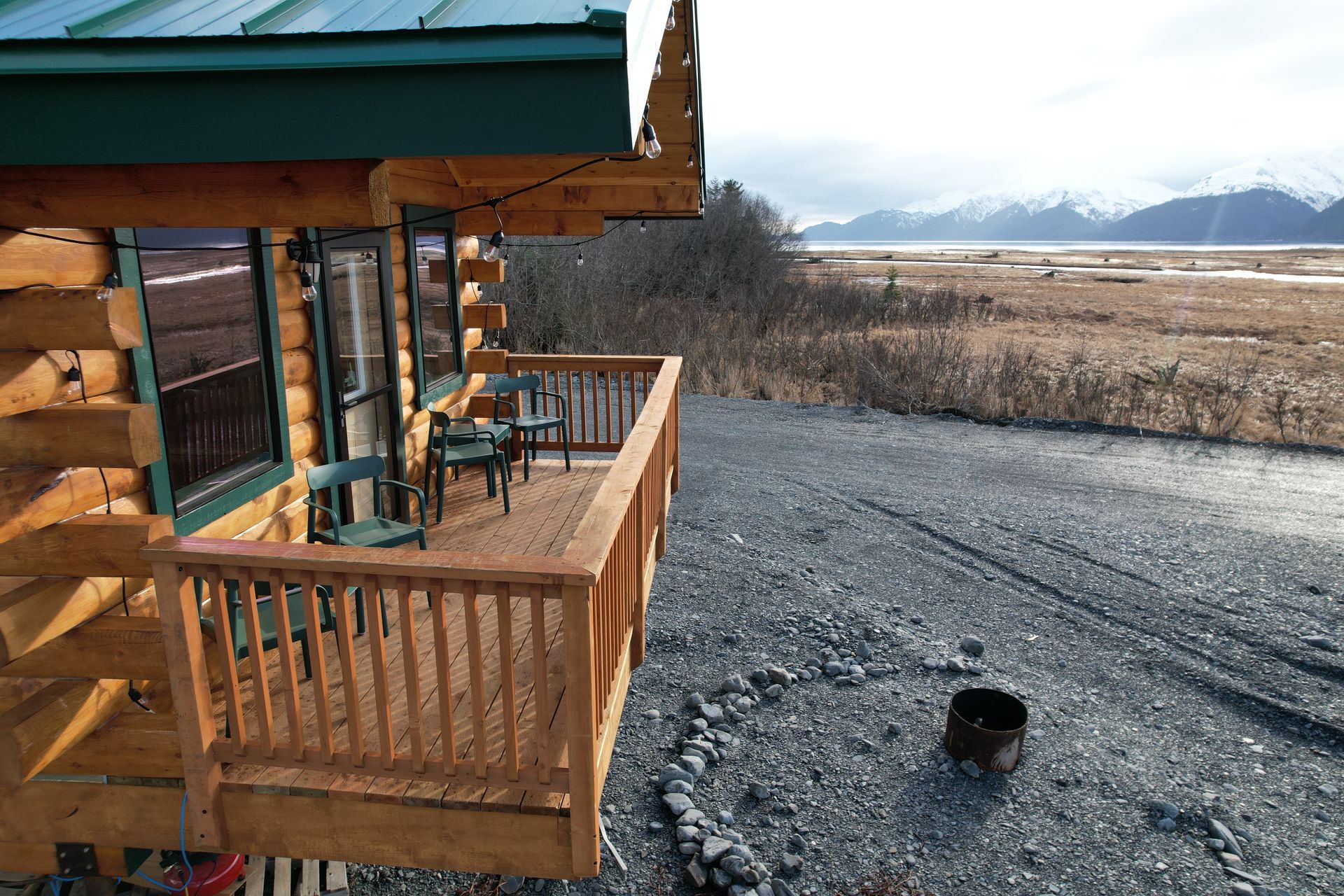 Log cabin with small deck overlooking a marsh and mountains. Gravel driveway, fire pit, green roof.