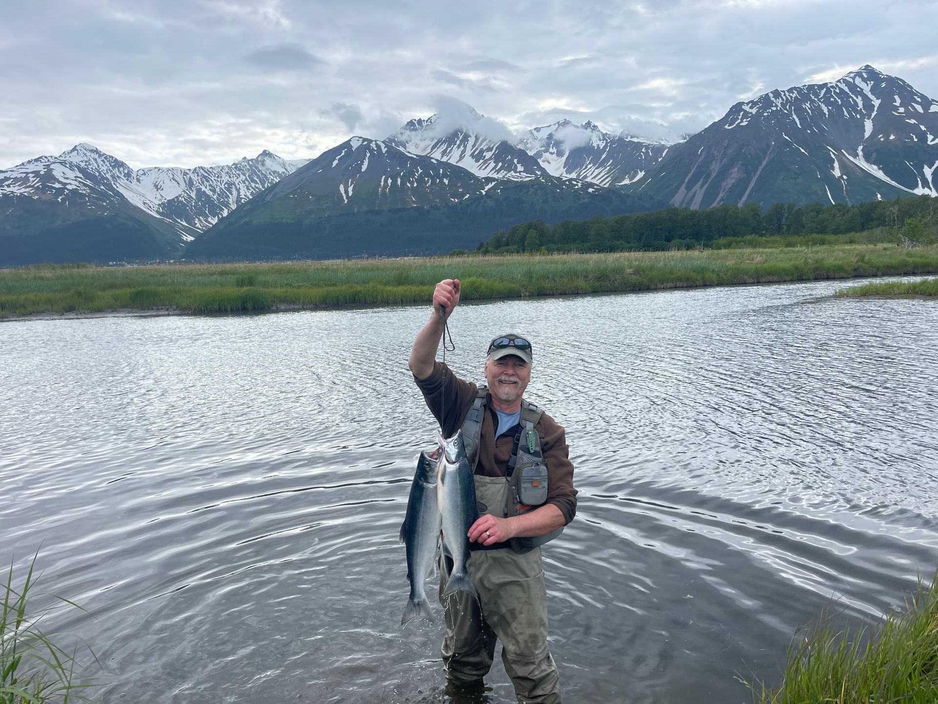Man in waders holds two fish, standing in water with mountains in background.