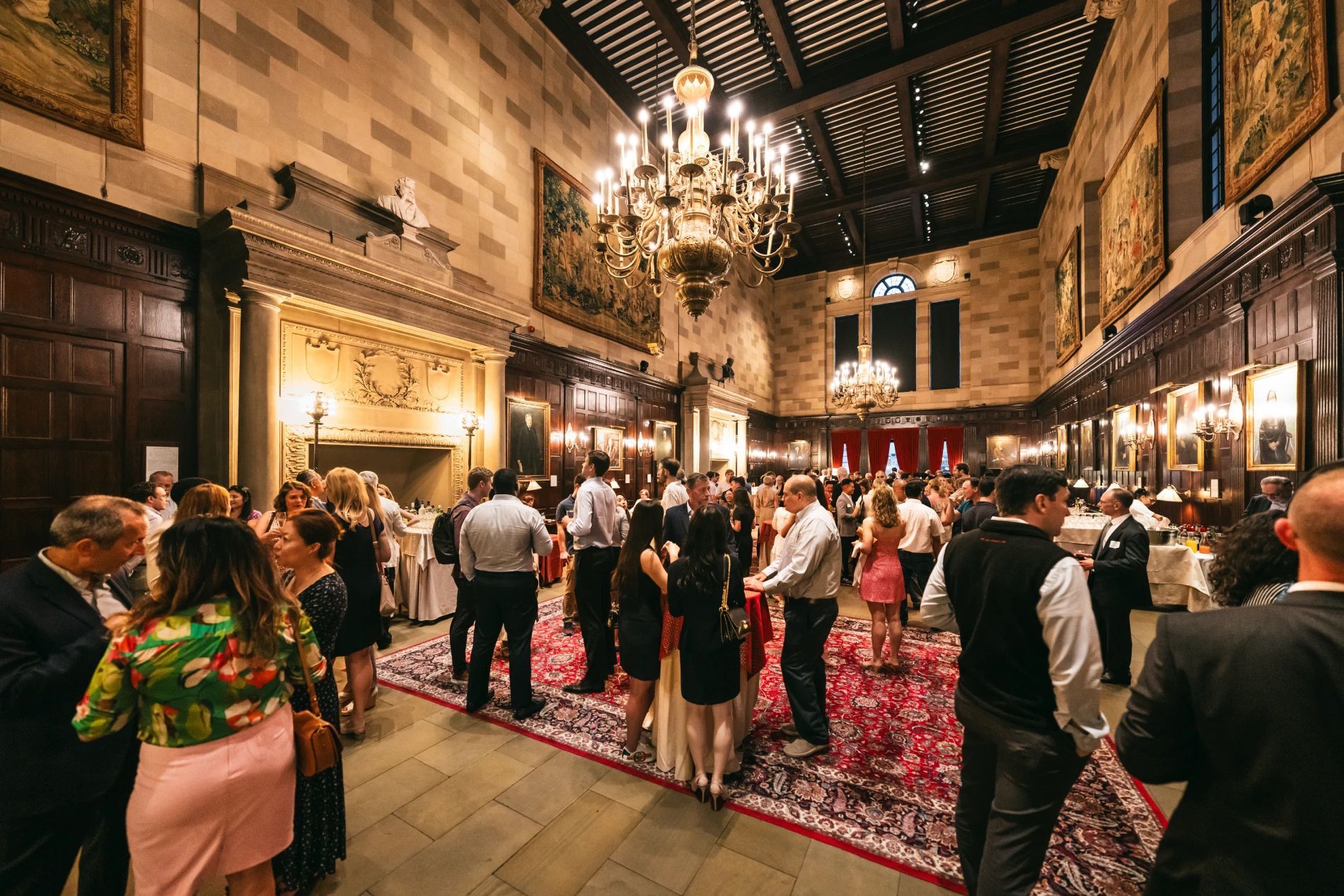 Formal event in a large room with a chandelier, people mingling, stone walls, and a patterned rug.