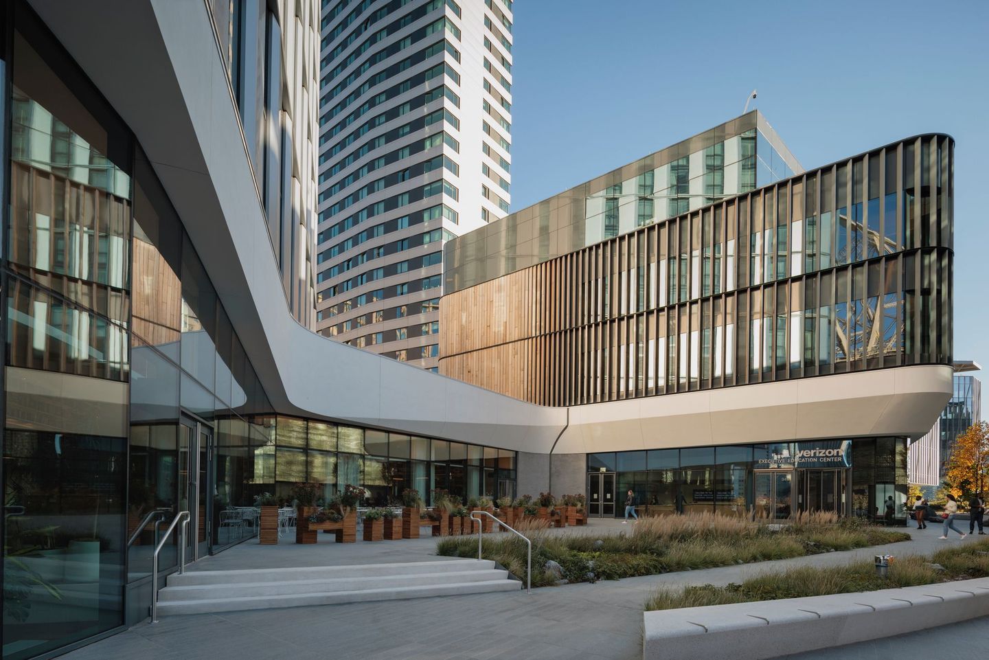 Modern building exterior with glass and wooden facade, outdoor seating, and a tall tower in the background.