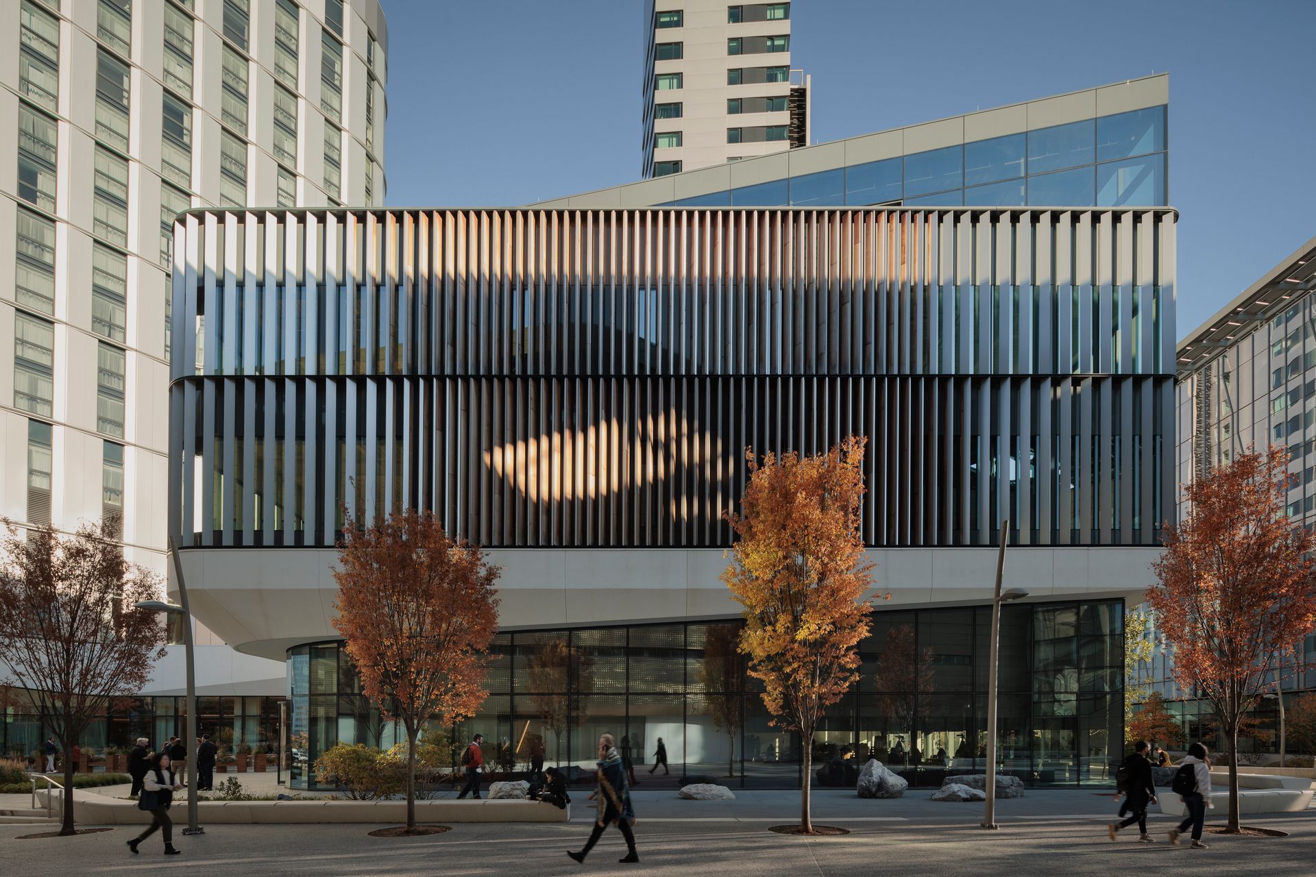 Modern building with vertical fins; people walk by trees with fall foliage.