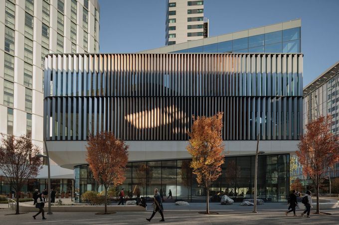 Modern building with vertical fins; people walk by trees with fall foliage.