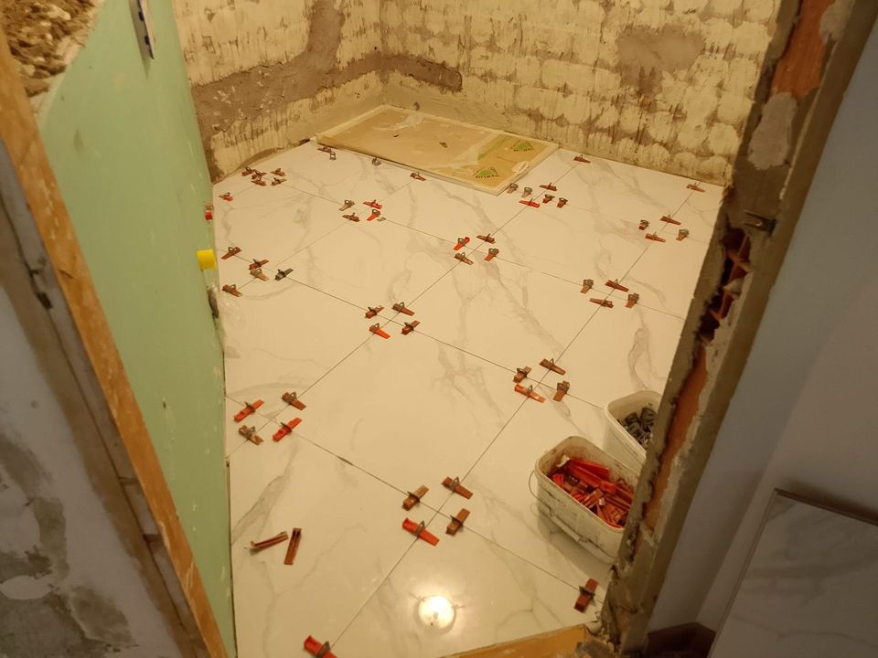 Piccola stanza piastrellata in fase di ristrutturazione con pavimento bianco decorato, pareti non rifinite e un vassoio per la vernice in un angolo.