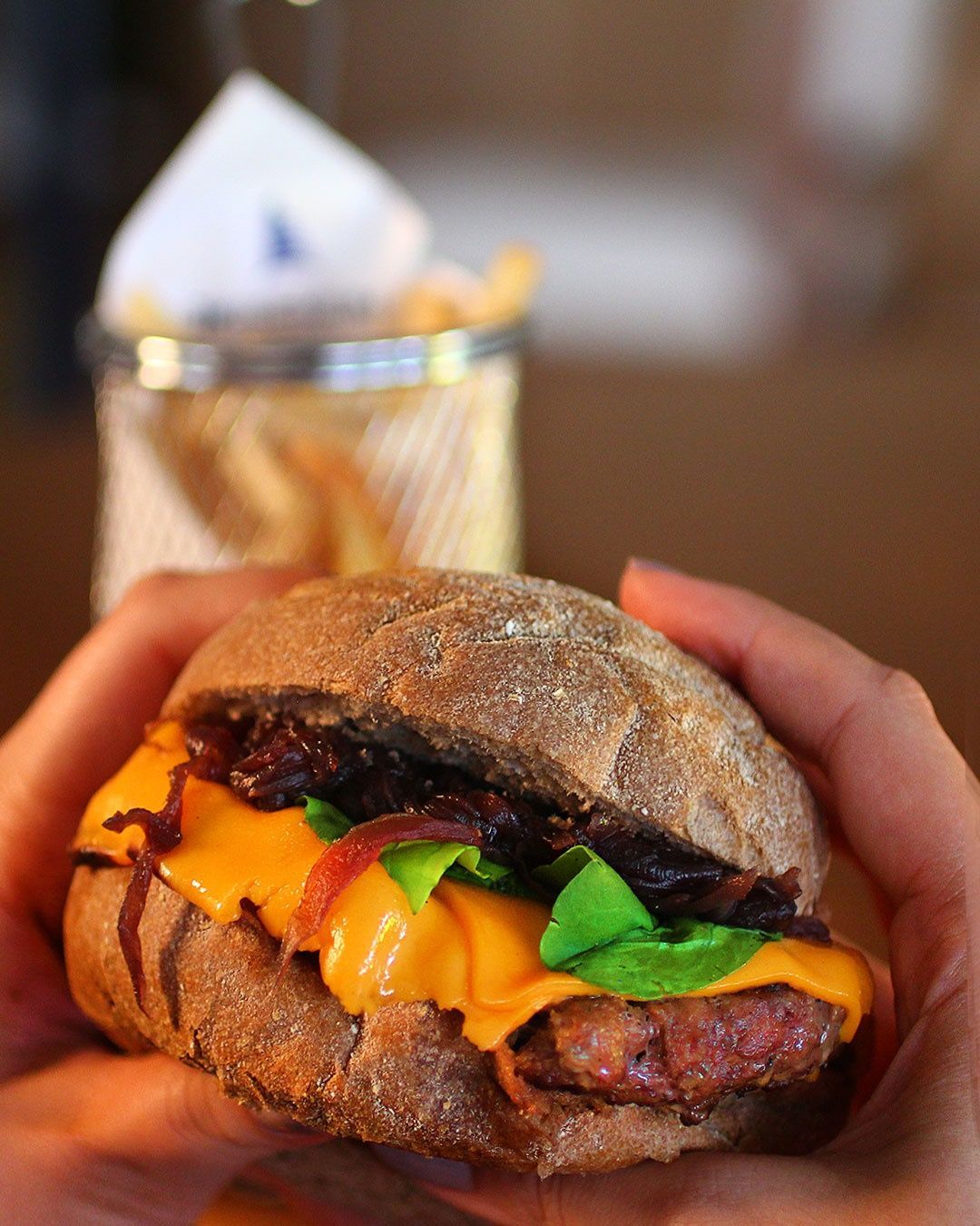 Pessoa segurando um hambúrguer com queijo, verduras e cebolas caramelizadas em um pão escuro; batatas fritas em uma cesta de metal atrás.