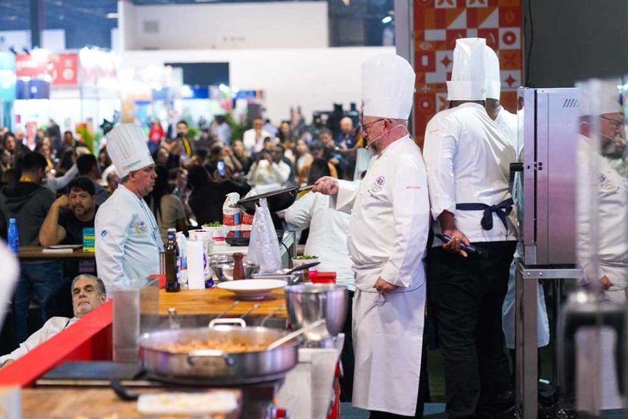 Chefs cozinhando em um evento culinário, interagindo em um fogão com uma multidão ao fundo.