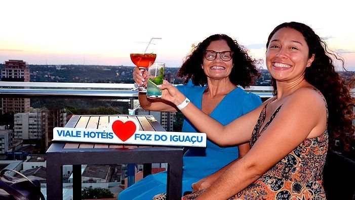 Duas mulheres em um terraço, brindando com coquetéis, sorrindo. Placa de Foz do Iguaçu, pôr do sol.