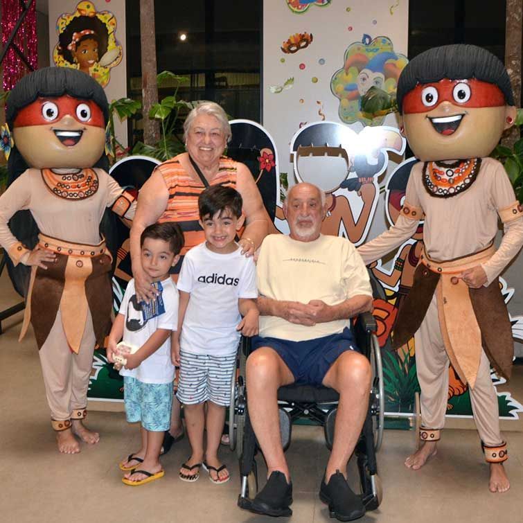 Família com dois mascotes fantasiados, posando para foto. Um idoso em uma cadeira de rodas sorri enquanto dois meninos e uma mulher estão ao seu redor.