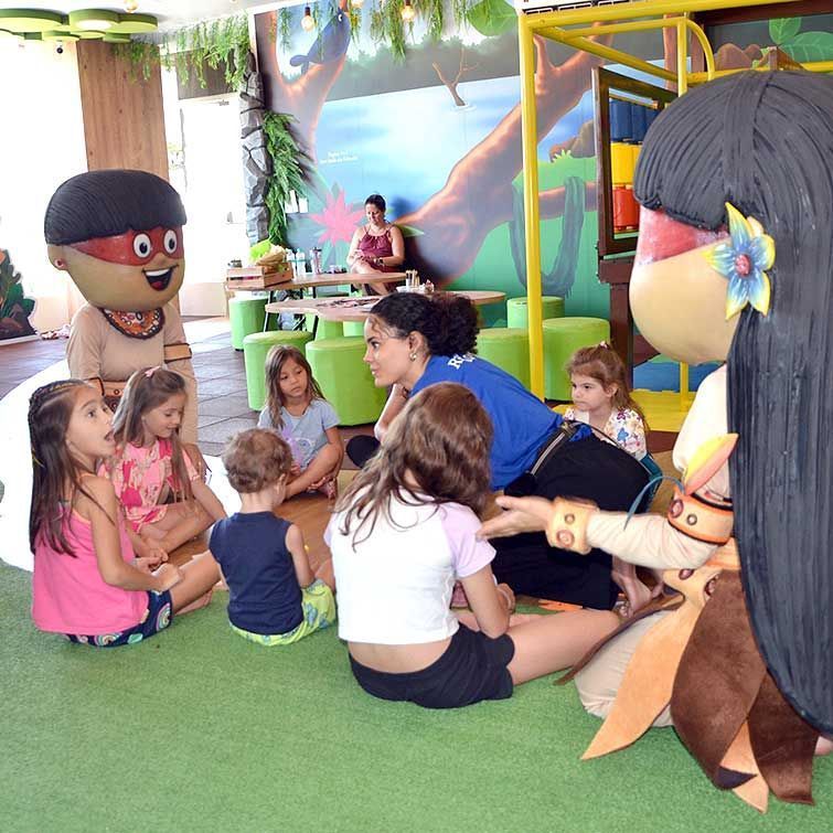 Crianças sentam-se em chão verde, ouvindo dois personagens fantasiados em uma área de recreação com um mural de selva.