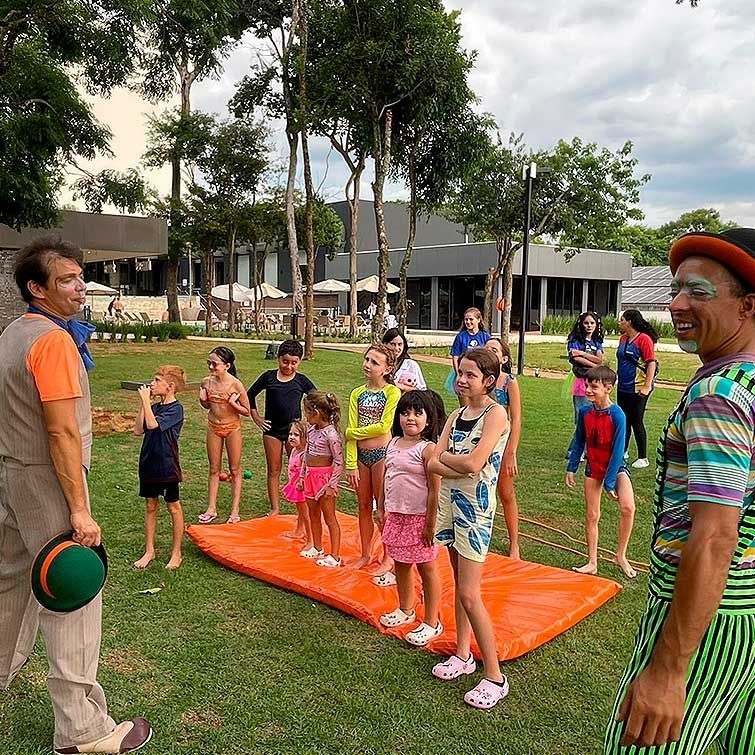 Dois palhaços com crianças em um tapete laranja em uma área gramada. As crianças usam trajes de banho; um prédio ao fundo.