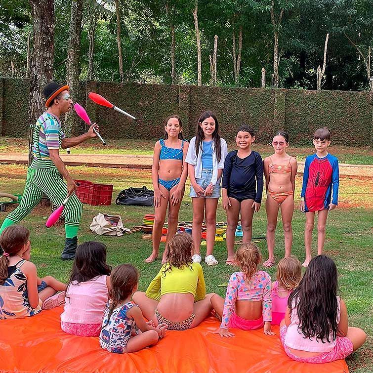 Palhaço fazendo malabarismo para crianças ao ar livre. Crianças assistem, algumas em trajes de banho. Grama verde, dia ensolarado.