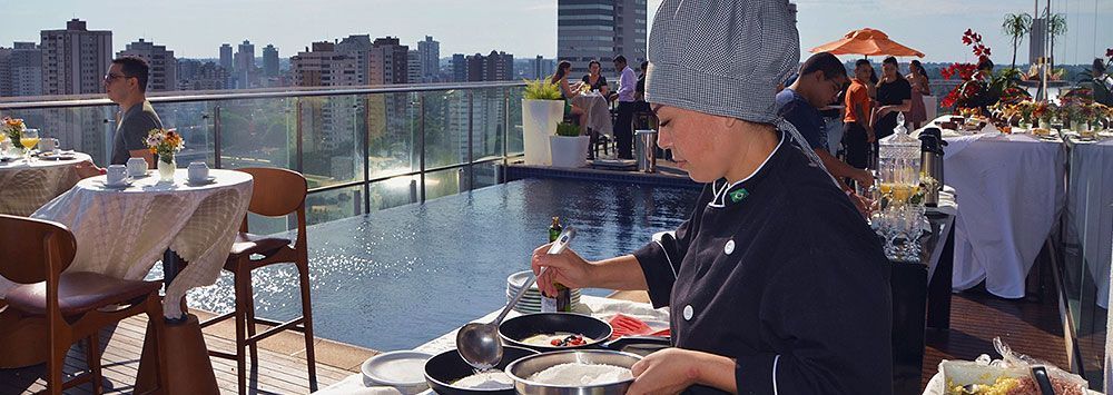 Chef cozinhando ao ar livre, perto de uma piscina, com vista para a cidade.