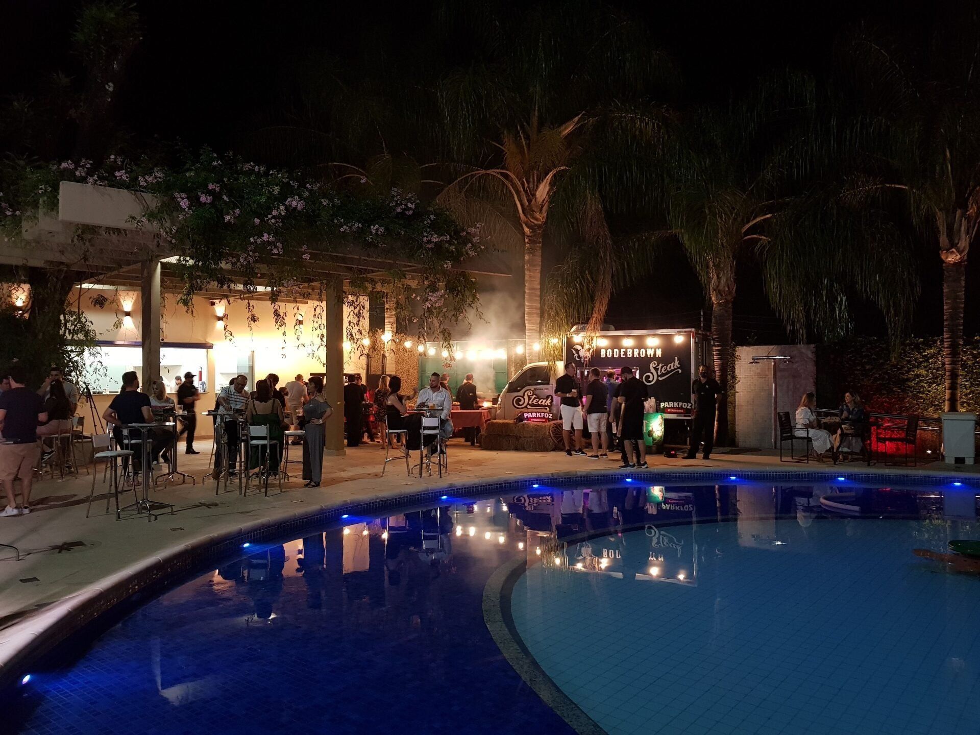 Festa noturna na piscina com pessoas, luzes, bar e palmeiras.