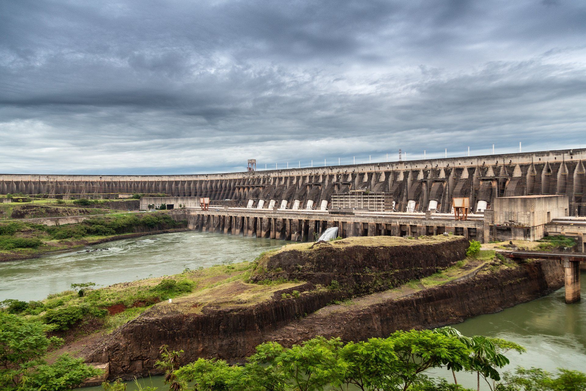 Uma grande barragem hidrelétrica em um rio sob um céu nublado; estrutura de concreto com água fluindo.