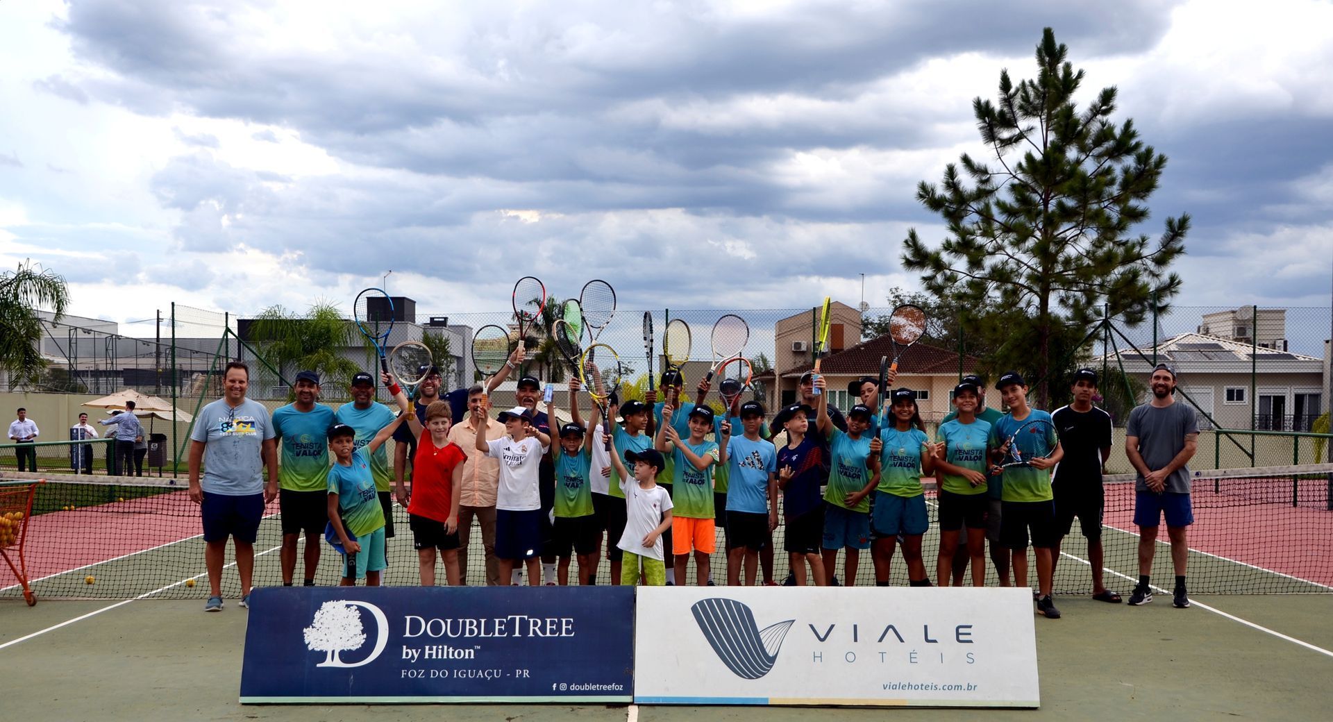 Grupo de tenistas comemorando em uma quadra sob um céu nublado. Jogadores seguram raquetes e faixas em primeiro plano.