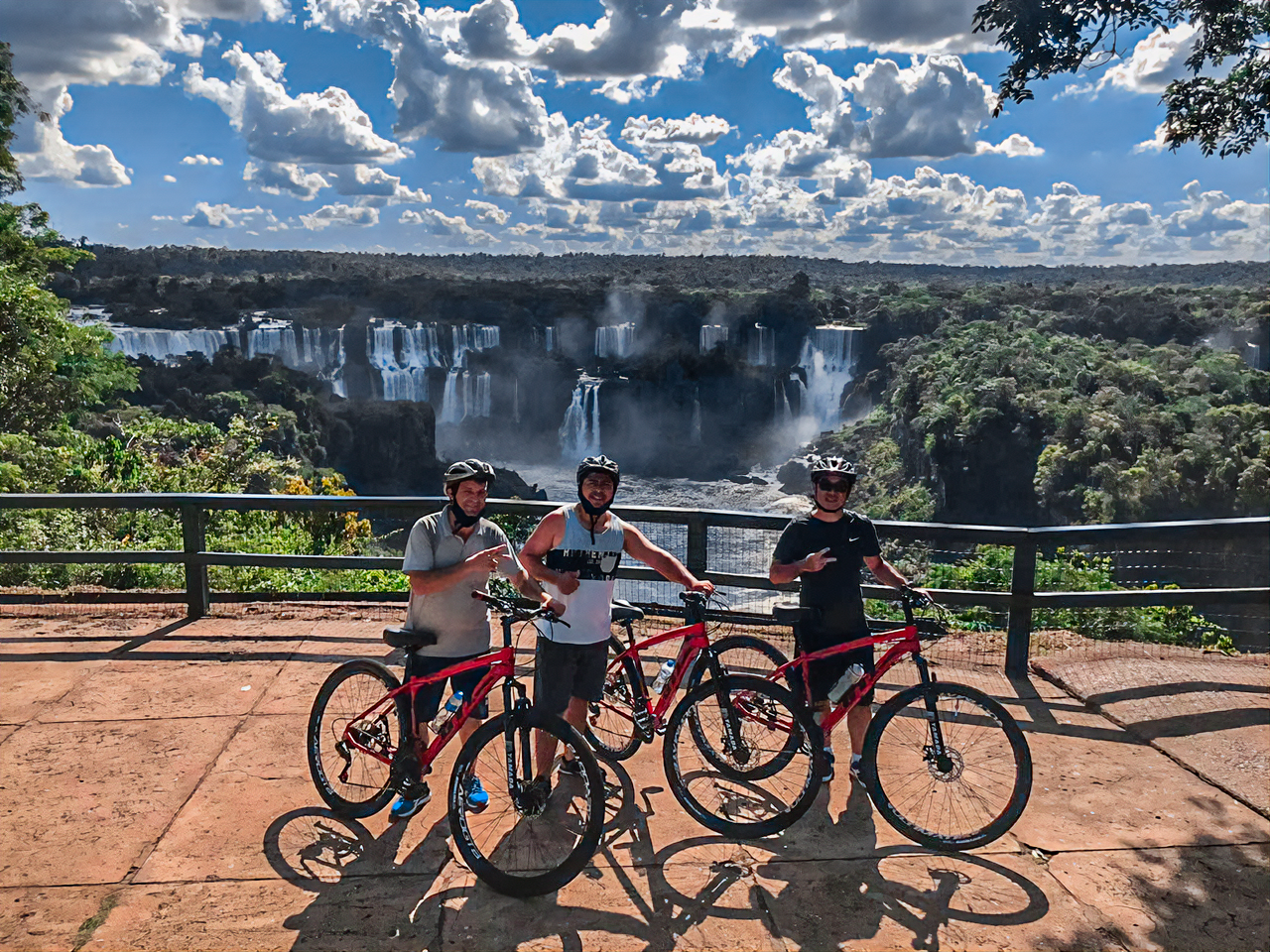 Três pessoas em bicicletas posam em frente às Cataratas do Iguaçu. Dia ensolarado.