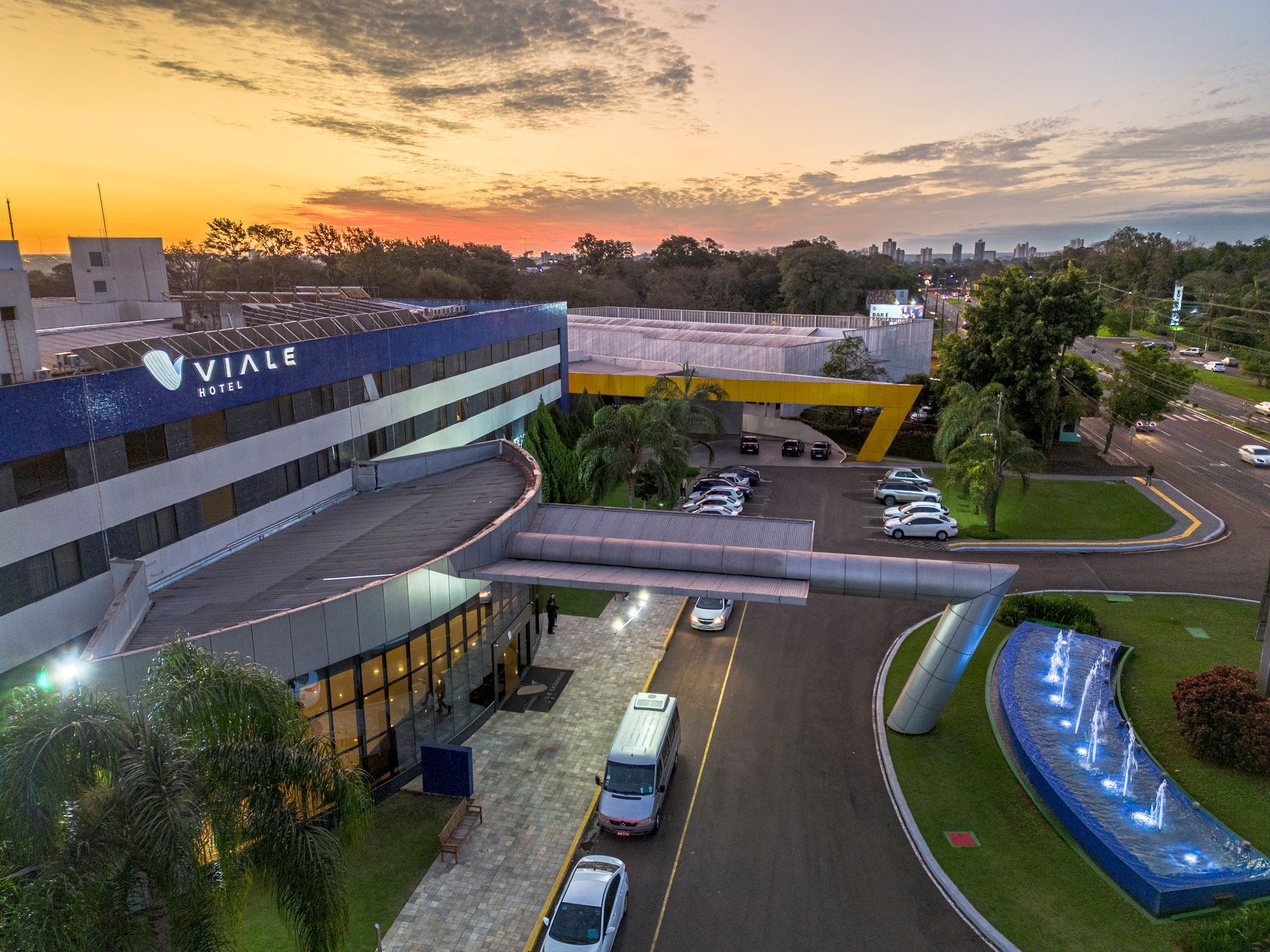 Fachada do hotel com detalhes em azul e amarelo, carros estacionados, ao pôr do sol.