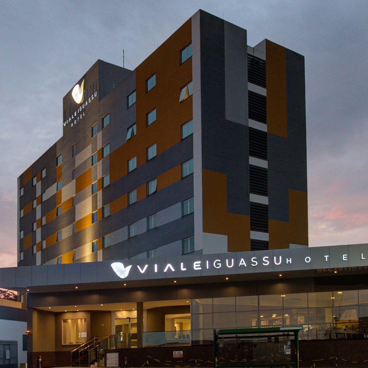 Fachada do Hotel Viale Iguassu ao entardecer. Prédio cinza e laranja com letreiro luminoso.