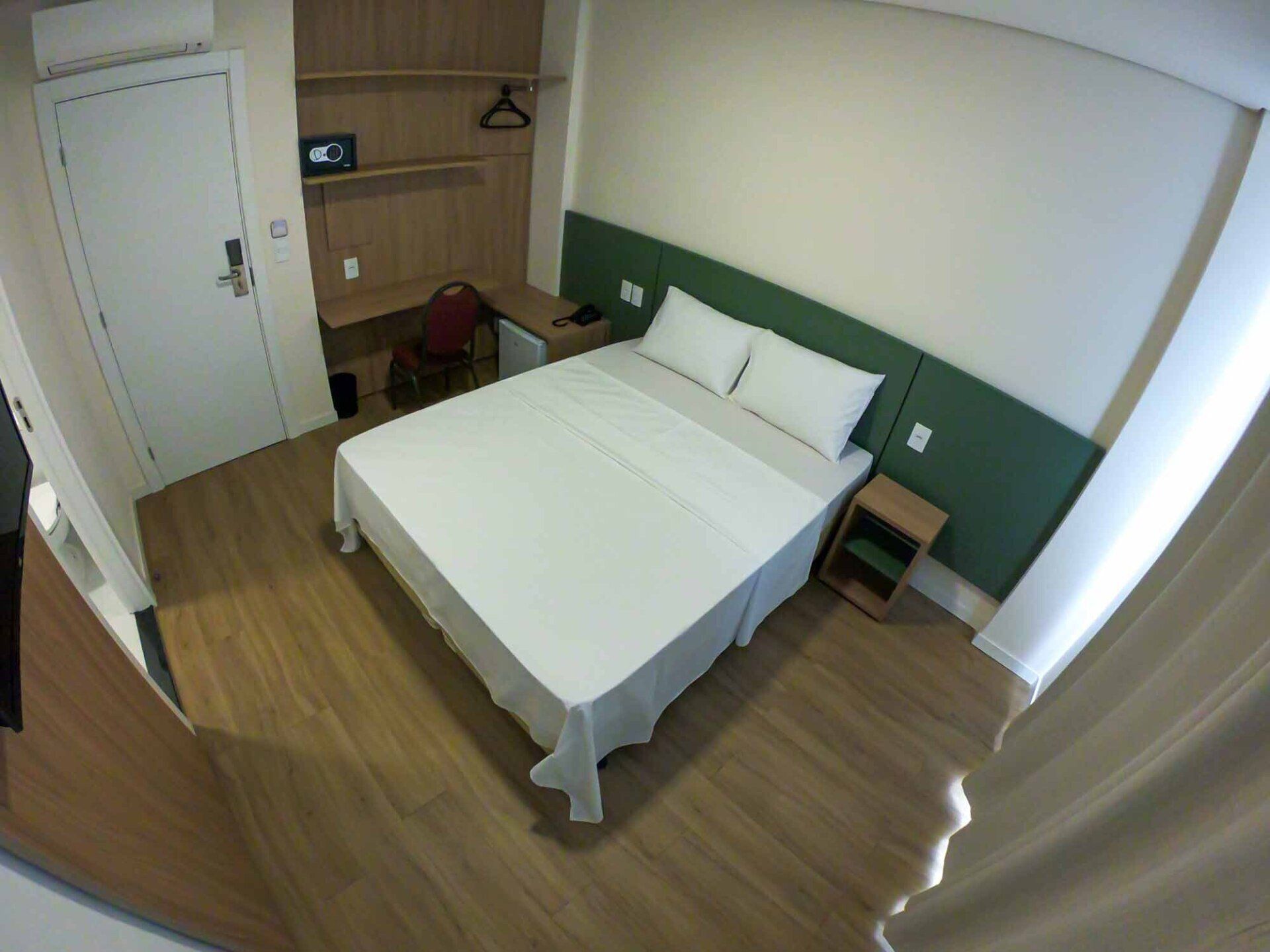 Quarto de hotel com cama branca, piso de madeira, escrivaninha e cabeceira verde.