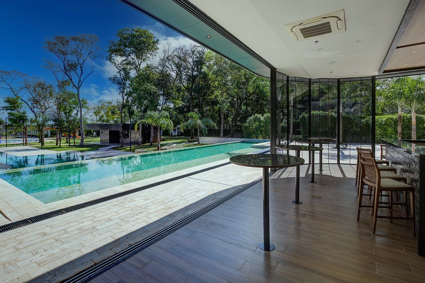 Um quarto moderno com vista para a piscina: paredes de vidro, bancos de bar, mesas, piscina, árvores, dia ensolarado.