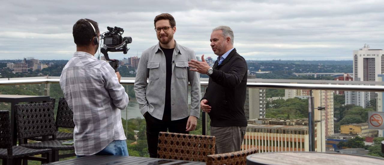 Três homens em um telhado, um filmando os outros dois. Céu nublado, horizonte da cidade ao fundo.