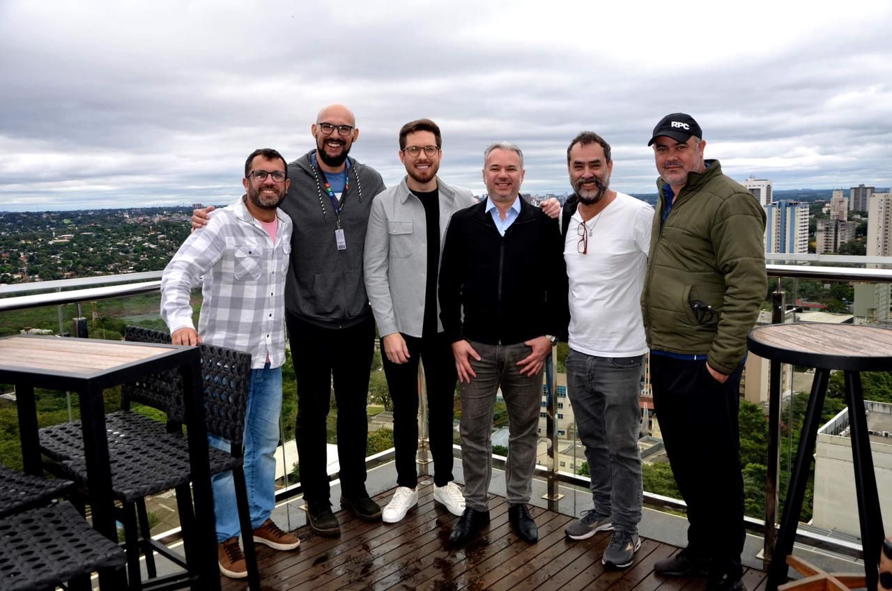 Seis homens posam em um terraço com vista para a paisagem urbana. Céu nublado. Alguns usam jaquetas e trajes casuais.