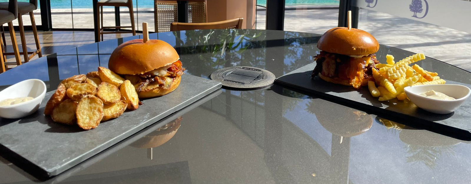 Dois hambúrgueres, batatas fritas e molhos em pratos retangulares. Um hambúrguer tem batatas rústicas, o outro tem batatas fritas.