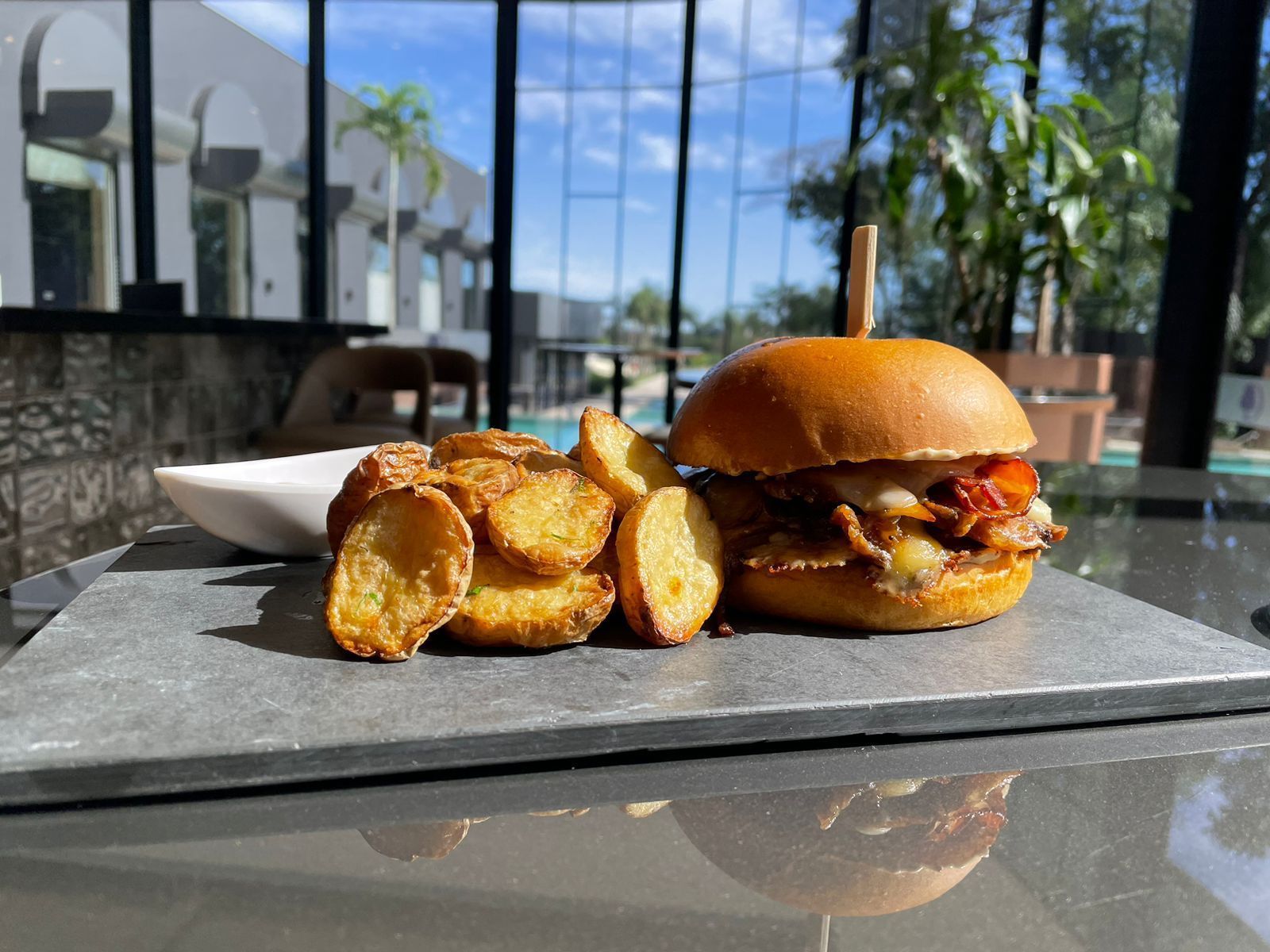 Hambúrguer com batatas fritas em um prato de ardósia, ao lado de uma tigela de molho. Ambiente externo com um prédio moderno ao fundo.