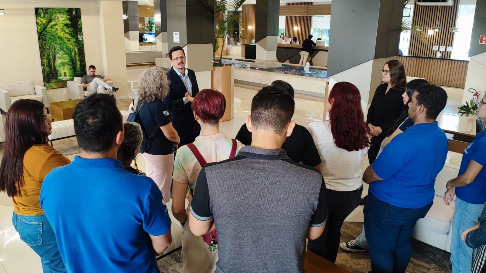 Grupo de pessoas ouvindo um homem de terno, em ambiente interno. Lobby de hotel com decoração moderna e arte.