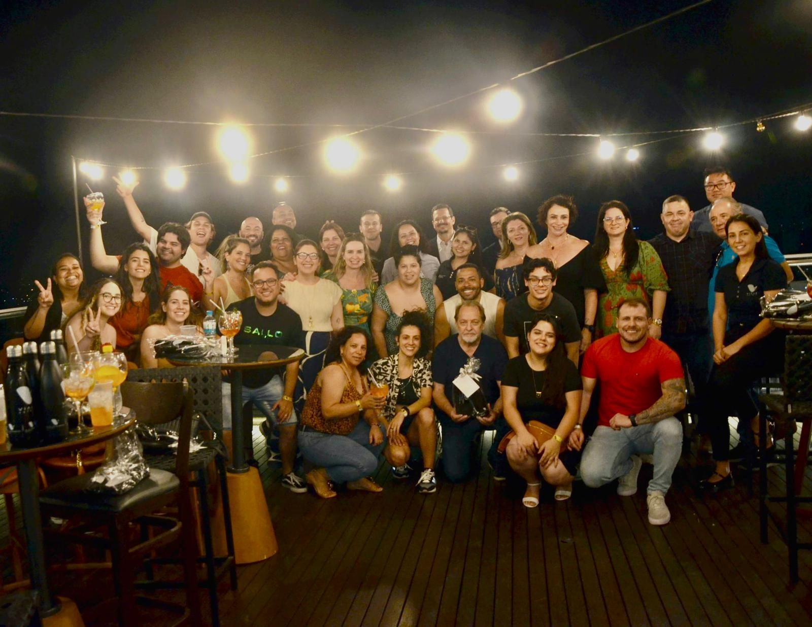 Grupo de pessoas em uma reunião ao ar livre à noite, sob luzes de corda, sorrindo.