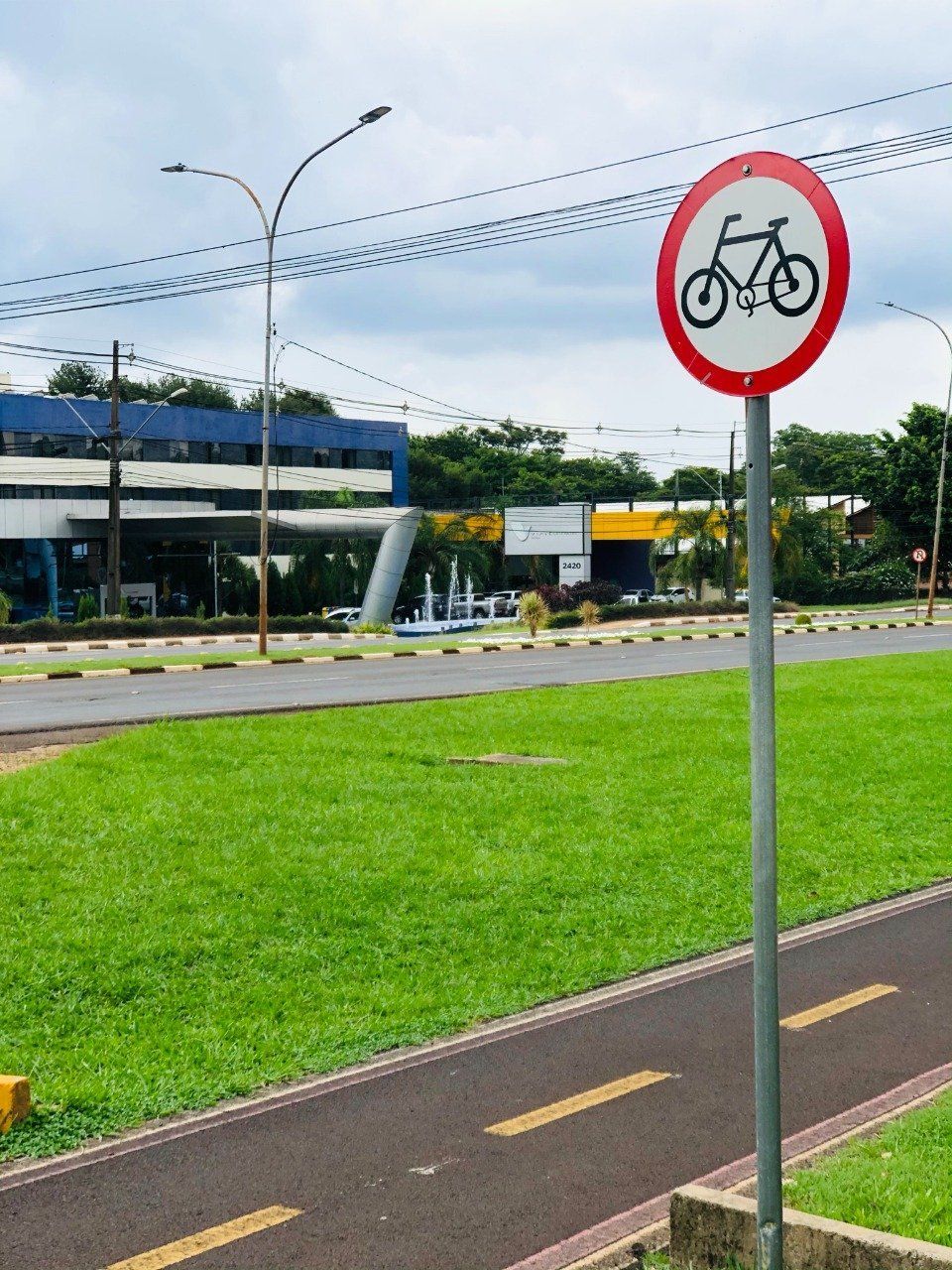 Placa de ciclovia com um círculo vermelho, ao lado de grama verde e uma estrada escura. Prédio azul ao fundo.