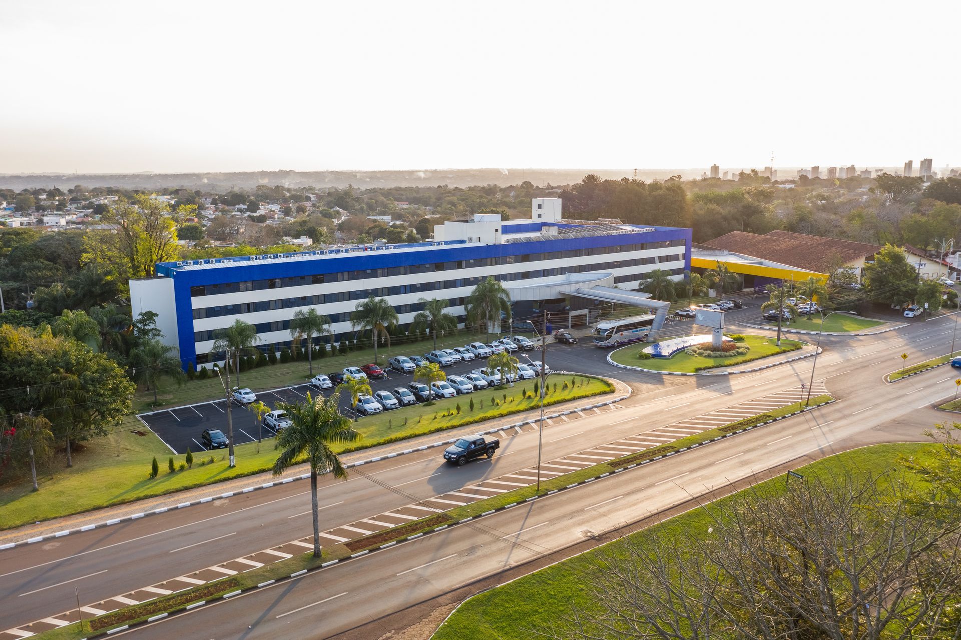 Grande edifício azul e branco com entrada circular para veículos e área de estacionamento, em um dia ensolarado.