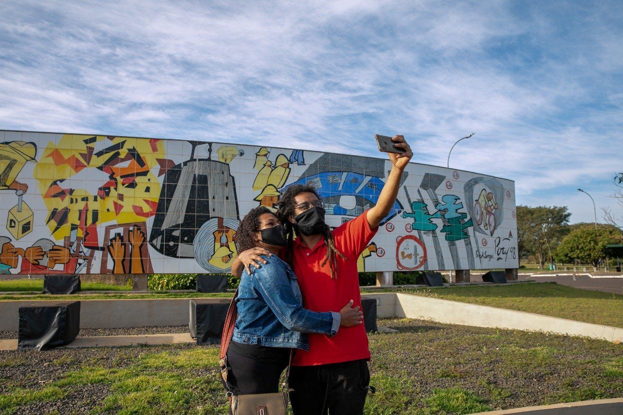 Duas pessoas com máscaras tiram uma selfie em frente a um mural colorido ao ar livre.