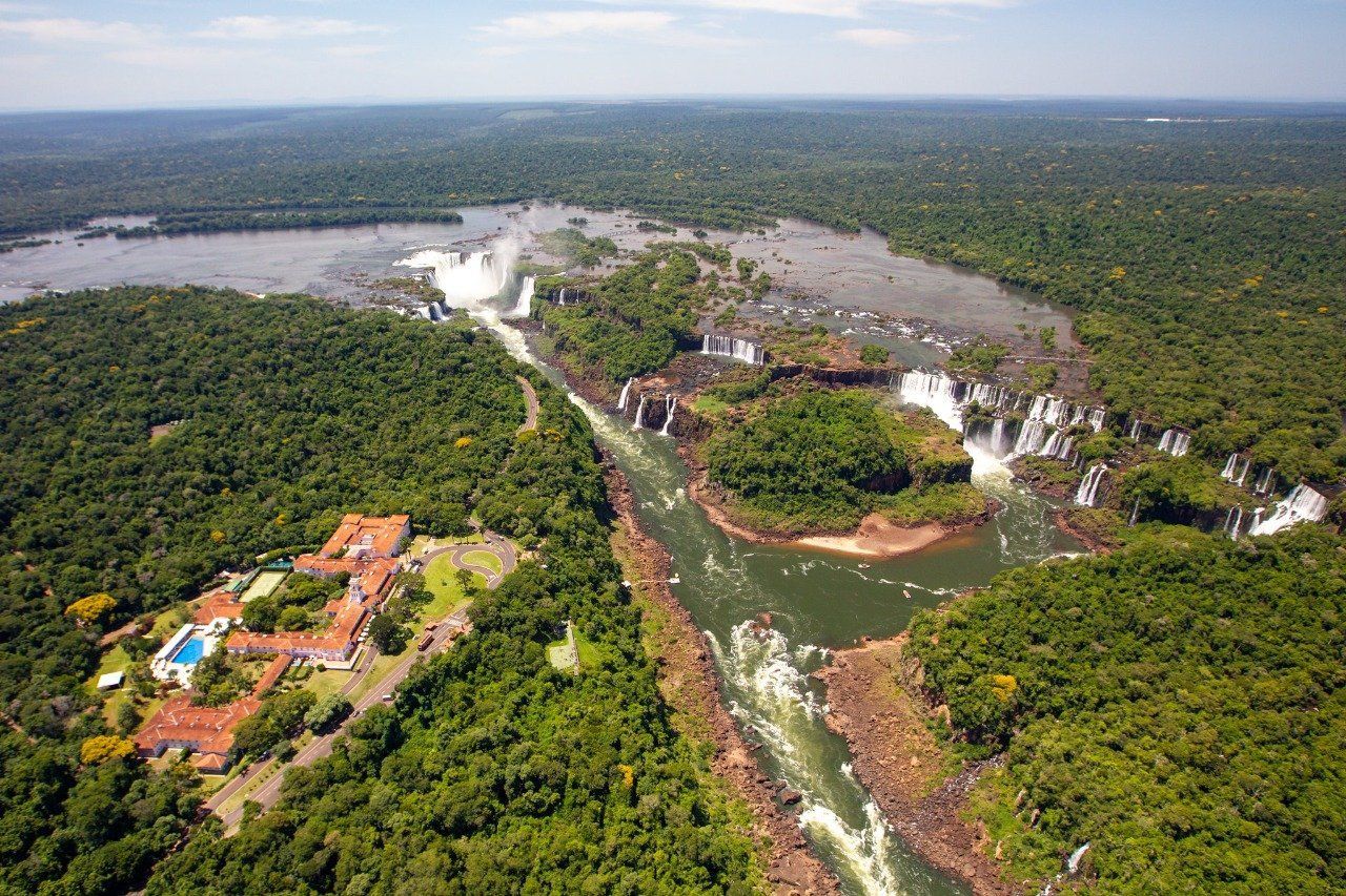 Vista aérea das Cataratas do Iguaçu, uma floresta verdejante cercada por cachoeiras e um hotel.
