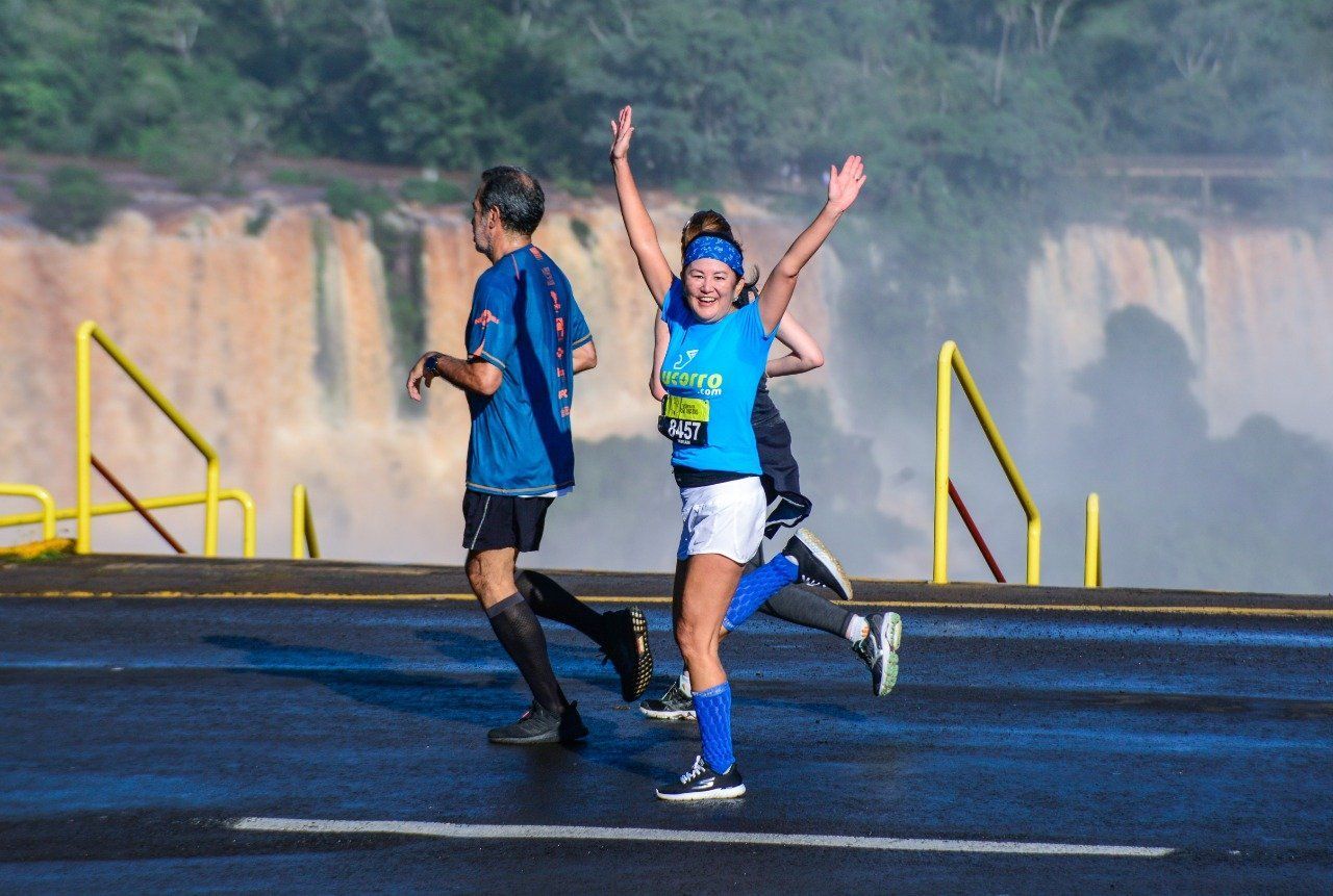 Mulher correndo com os braços levantados, roupa azul, em frente a cachoeiras.