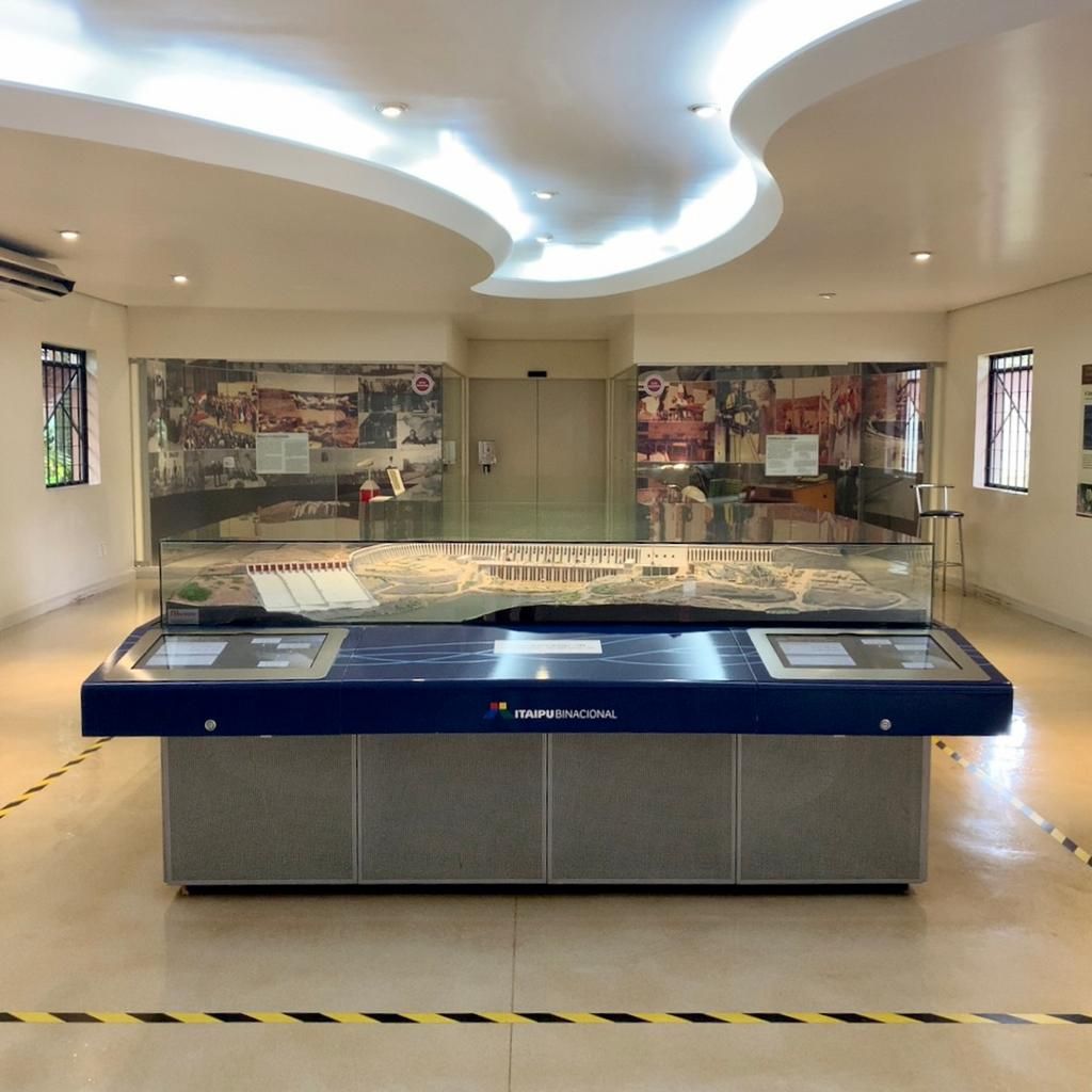 Sala de exposições do museu. Expositor azul com vitrine de vidro contendo uma maquete. Grandes vitrines de vidro na parede do fundo. Interior bege, luz natural.